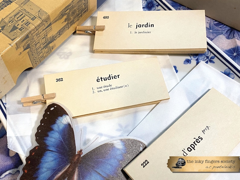 May include: A set of flashcards with French vocabulary words and definitions. The cards are beige with black text and are held together with wooden clothespins. The cards are on a white surface with a blue and white patterned background.
