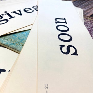 May include: Close-up of a stack of cards with the word "SOON" printed in black on a cream-colored background.