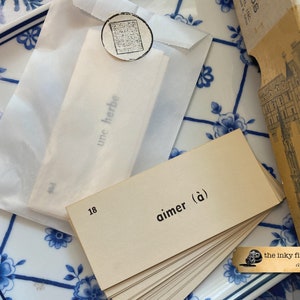 May include: A stack of beige index cards with the word "aimer (&agrave;)" printed on the top card. The cards are on a blue and white patterned surface. The text "the inky fingers society a.c.pantelakis" is printed on a small black and white sticker in the lower right corner of the image.