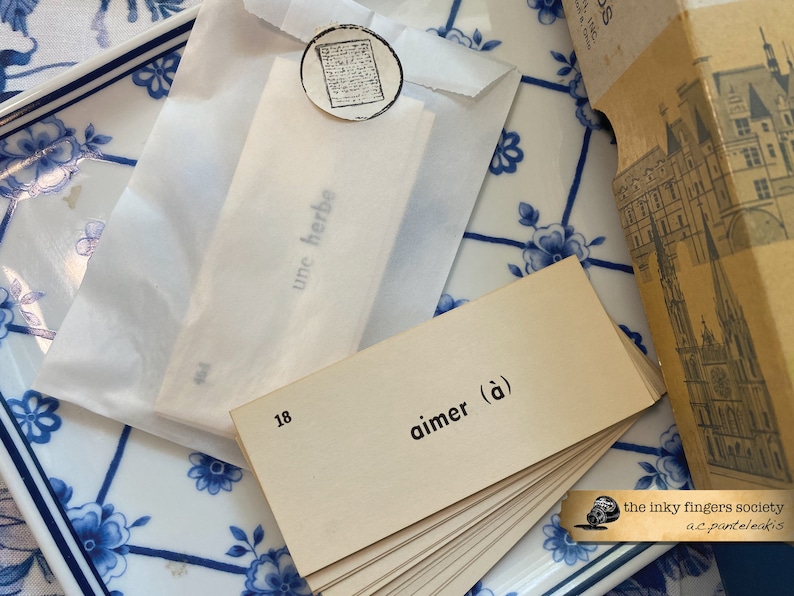May include: A stack of beige index cards with the word "aimer (&agrave;)" printed on the top card. The cards are on a blue and white patterned surface. The text "the inky fingers society a.c.pantelakis" is printed on a small black and white sticker in the lower right corner of the image.