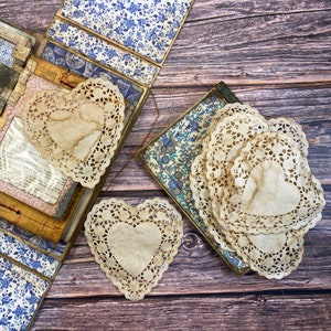 Può includere: Tre delicati tovaglioli di carta a forma di cuore, con intricati motivi di pizzo, sono esposti su una superficie di legno. I tovaglioli sono di colore beige chiaro e hanno un'estetica vintage.