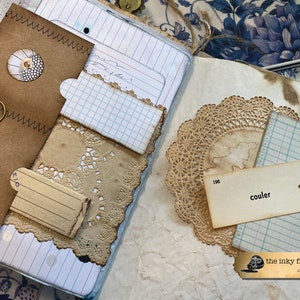 May include: A vintage journal with brown paper covers, a paper doily, and a tag that says "couler". The journal has a worn and aged look, with visible stitching and a paper clip.