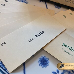 May include: A stack of beige index cards with black numbers and text. The top card has the French phrase "une herbe" written on it. The bottom card has the word "garden" written in green text with the word "gardener" underneath.