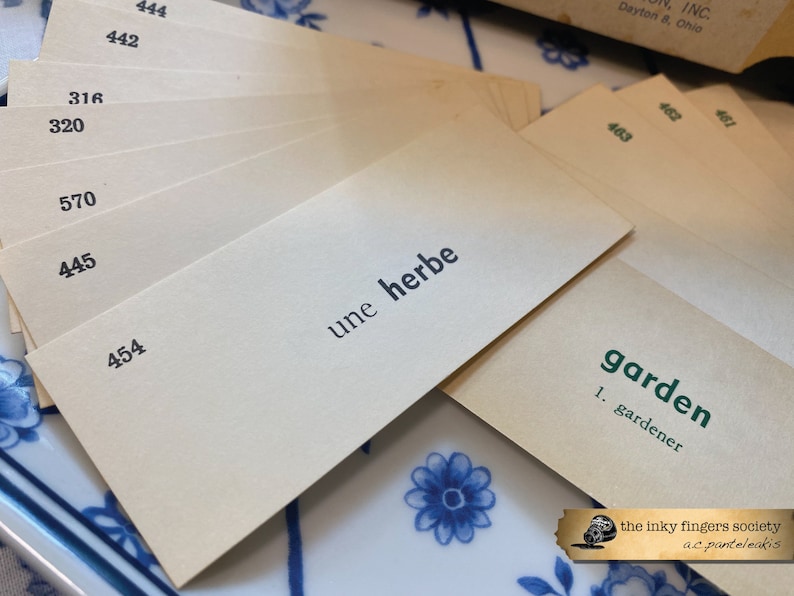 May include: A stack of beige index cards with black numbers and text. The top card has the French phrase "une herbe" written on it. The bottom card has the word "garden" written in green text with the word "gardener" underneath.