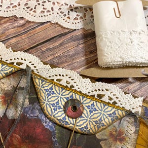 May include: Close-up of craft supplies including white lace trim, a roll of lace-edged paper, and a decorative paper pocket with a floral and blue tile pattern. The items are arranged on a wooden surface.