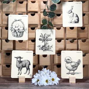 May include: Five Easter-themed cards with black and white illustrations. The cards are displayed on a wooden shelf with small drawers. The illustrations include a basket of eggs, a cross with flowers, a lamb, a chick, and a rabbit.