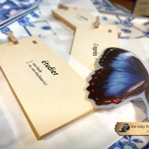 May include: A set of beige index cards with the French word "&eacute;tudier" written on one card. The word is defined as "to study" in French. The cards are held together with a wooden clothespin. A blue butterfly sticker is attached to the cards.
