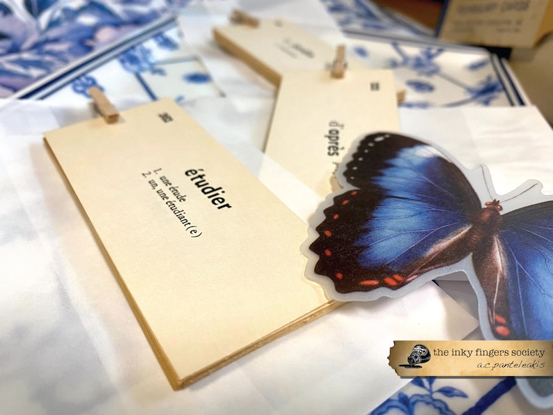 May include: A set of beige index cards with the French word "&eacute;tudier" written on one card. The word is defined as "to study" in French. The cards are held together with a wooden clothespin. A blue butterfly sticker is attached to the cards.