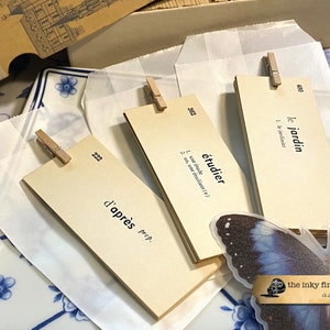 May include: Three beige index cards with French vocabulary words and definitions. The words are "le jardin", "&eacute;tudier", and "d'apr&egrave;s". The cards are clipped to white paper envelopes with wooden clothespins. A blue butterfly sticker is on the bottom right corner of the image.