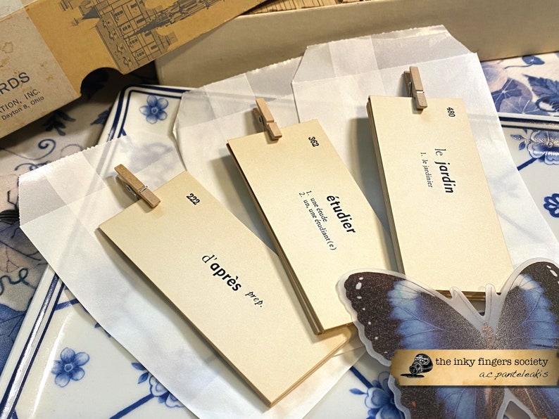 May include: Three beige index cards with French vocabulary words and definitions. The words are "le jardin", "&eacute;tudier", and "d'apr&egrave;s". The cards are clipped to white paper envelopes with wooden clothespins. A blue butterfly sticker is on the bottom right corner of the image.