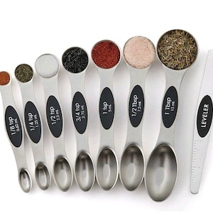 May include: A set of stainless steel measuring spoons and a leveler. Each spoon has a black oval with white text indicating the measurement in teaspoons and tablespoons, and milliliters. The spoons are filled with various spices.