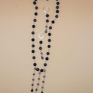 Pode incluir: Um conjunto de colar e brincos com contas redondas escuras e pingentes em forma de gota. O colar tem um design longo e em cascata com vários fios. Os brincos são argolas simples. A joia é feita com metal prateado.