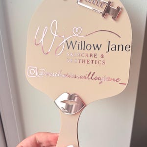 May include: A beige handheld mirror featuring rose gold accents, including a syringe icon at the top and a lip icon near the handle. The text reads Willow Jane SKINCARE & AESTHETICS with an Instagram icon and the handle @aesthetics.willowjane_.