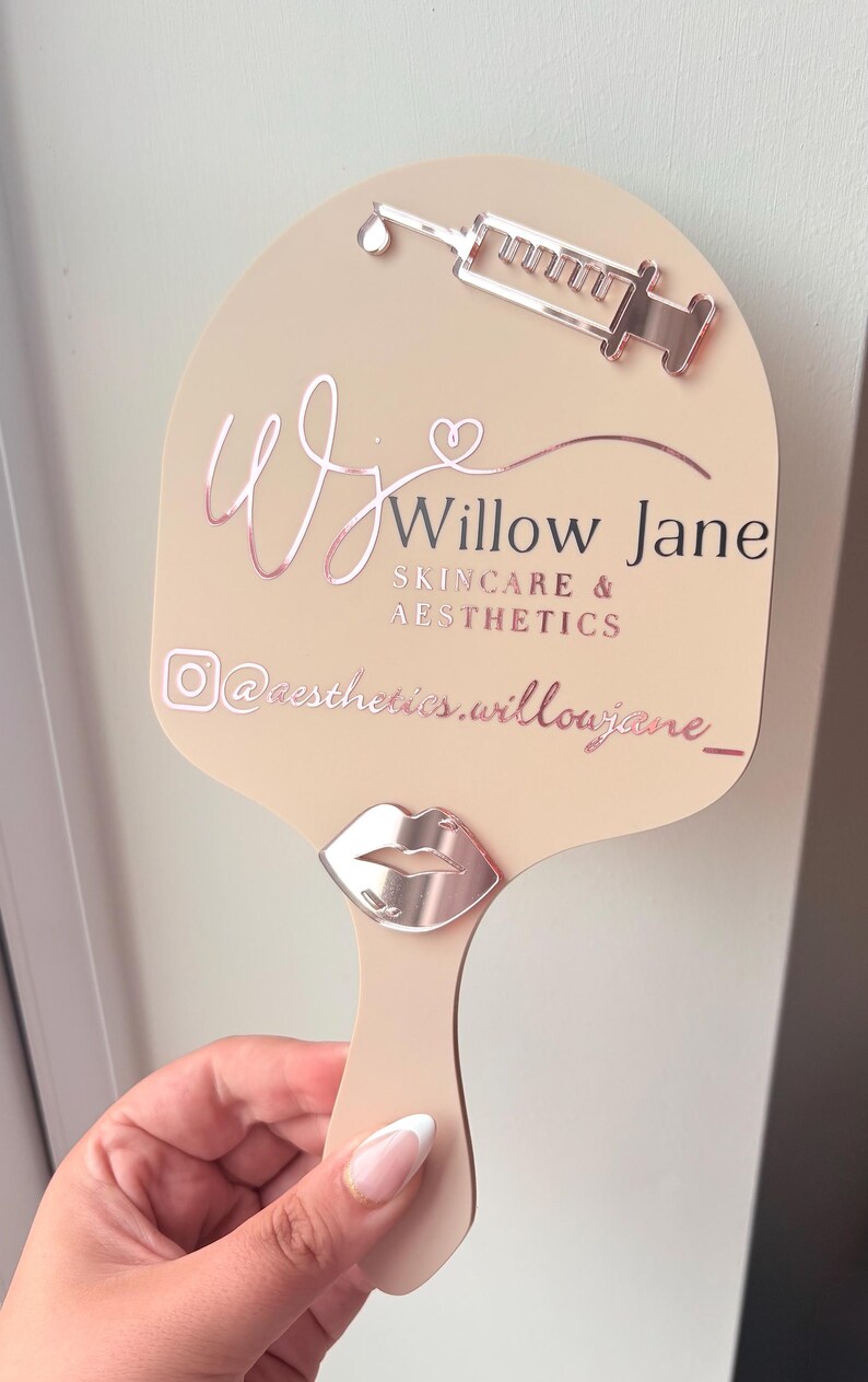 May include: A beige handheld mirror featuring rose gold accents, including a syringe icon at the top and a lip icon near the handle. The text reads Willow Jane SKINCARE & AESTHETICS with an Instagram icon and the handle @aesthetics.willowjane_.