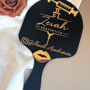 May include: A black handheld mirror featuring a gold-colored syringe icon and the text Zeiah AESTHETICS. Below the text is an Instagram icon followed by the text @Zeiah Aesthetics and a gold-colored lip icon at the base of the mirror handle.