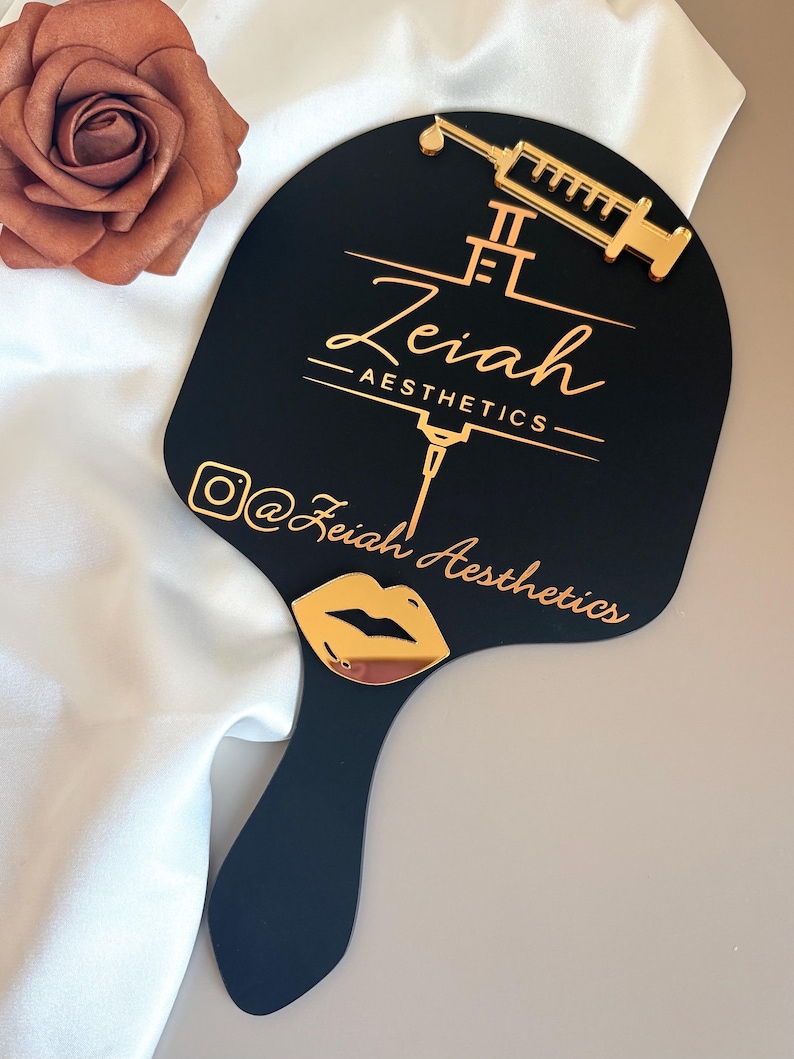May include: A black handheld mirror featuring a gold-colored syringe icon and the text Zeiah AESTHETICS. Below the text is an Instagram icon followed by the text @Zeiah Aesthetics and a gold-colored lip icon at the base of the mirror handle.