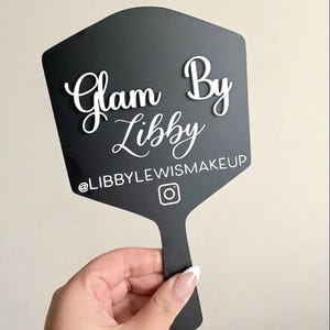 Może przedstawiać: Czarne sześciokątne lusterko ręczne z białym wypukłym napisem Glam By Libby czcionką skryptową, z białym nadrukiem @LIBBYLEWISMAKEUP oraz ikoną Instagrama umieszczonymi poniżej gł&oacute;wnej nazwy na powierzchni lusterka.