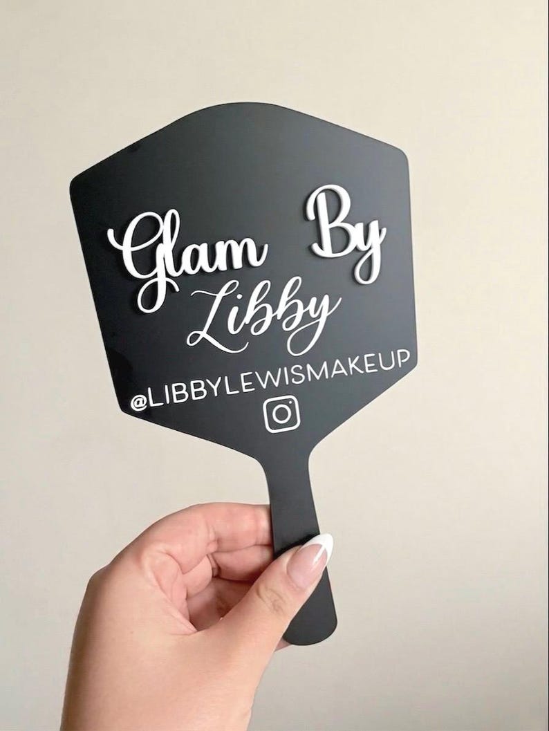 Może przedstawiać: Czarne sześciokątne lusterko ręczne z białym wypukłym napisem Glam By Libby czcionką skryptową, z białym nadrukiem @LIBBYLEWISMAKEUP oraz ikoną Instagrama umieszczonymi poniżej gł&oacute;wnej nazwy na powierzchni lusterka.