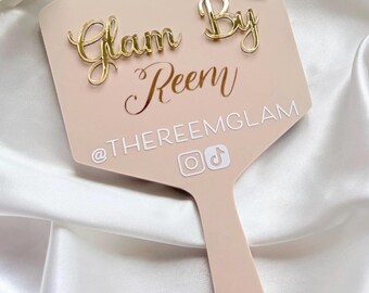 May include: A light pink handheld mirror featuring a gold acrylic lipstick icon and the gold script text Glam By Reem. Below the script, the white text @THEREEMGLAM is displayed alongside white Instagram and TikTok social media icons.