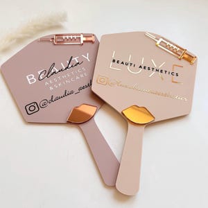 May include: Two blush-coloured hand mirrors with gold and rose gold accents. One mirror has the text "Claudia Aesthetics & Skincare" and the other reads "Luxe Beauti Aesthetics". Each has a lip-shaped handle.