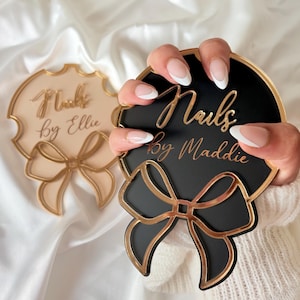 May include: Two circular acrylic signs with decorative bow accents at the bottom. One sign is black with gold lettering reading Nails by Maddie, and the other is beige with gold lettering reading Nails by Ellie. Both feature gold-toned edges.