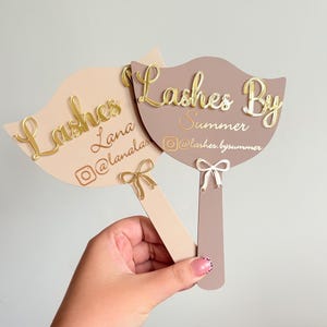May include: Two handheld fan-shaped signs in beige and mauve. The beige sign features gold text reading Lashes Lana and an Instagram handle. The mauve sign features gold text reading Lashes By Summer and an Instagram handle. Both have small bows.