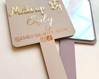 Könnte beinhalten: Handspiegel mit rechteckiger Spiegelfläche und Griff. Die Spiegelfläche ist hellgrau mit goldfarbenem Schriftzug "Makeup By Emily" und den Social-Media-Handles. Oben befindet sich ein goldenes Lippenstift-Design.