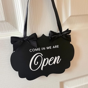 May include: A black scalloped-edge sign with white text that reads COME IN WE ARE Open. The sign features two black satin ribbon bows at the top corners and is suspended by a matching black ribbon for hanging, creating a clear and visible display.