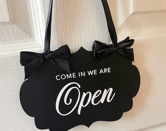May include: A black scalloped-edge hanging sign with white text that reads "COME IN WE ARE Open". The sign features two black satin ribbon bows at the top corners and is suspended by a matching black ribbon for display on a door or wall.