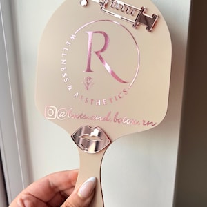 May include: A beige handheld mirror featuring a rose gold metallic syringe icon at the top and a rose gold metallic lip icon at the base. The center displays a rose gold circular logo with the letter R, the text Wellness & Aesthetics, and @boots.and.botox.rn.