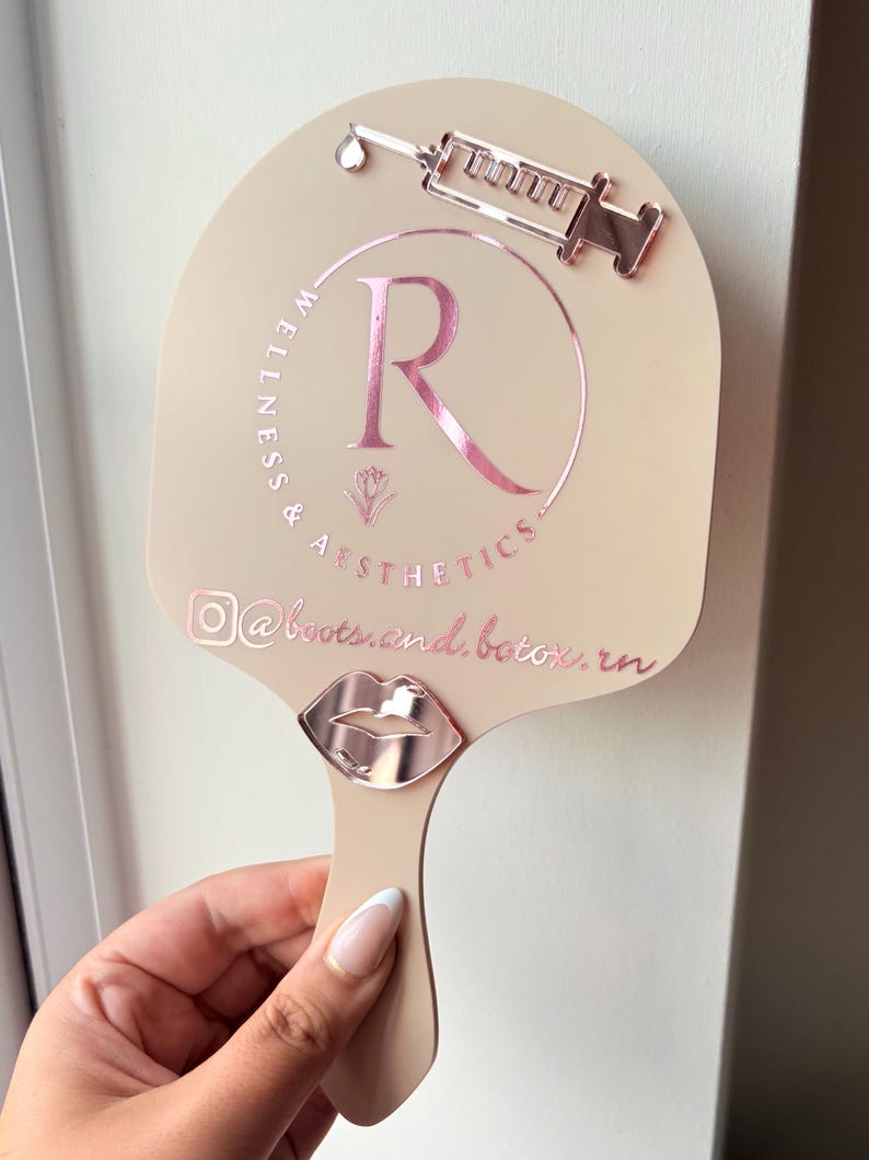 May include: A beige handheld mirror featuring a rose gold metallic syringe icon at the top and a rose gold metallic lip icon at the base. The center displays a rose gold circular logo with the letter R, the text Wellness & Aesthetics, and @boots.and.botox.rn.