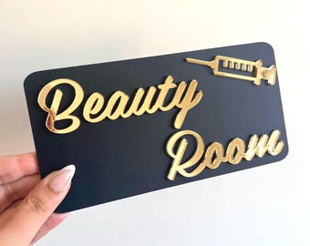 May include: A rectangular black sign featuring the gold-colored text Beauty Room in a cursive script font. A gold-colored syringe icon is positioned in the upper right corner of the sign. The sign has rounded corners and a smooth, matte finish.