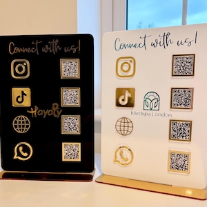 Könnte beinhalten: Zwei Acrylschilder mit goldenen Symbolen und QR-Codes für soziale Medien. Ein Schild ist schwarz mit dem Text "Connect with us!" und "Hayaty". Das andere Schild ist weiß mit dem Text "Connect with us!" und "Medspa London".
