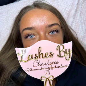 May include: A light pink handheld mirror with a scalloped top edge and a gold bow accent at the base. The mirror features the gold text Lashes By Charlotte, the handle text @blossombeautybycharlotte, and a small gold Instagram camera icon.