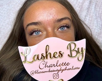 May include: A light pink handheld mirror with a scalloped top edge and a handle. The mirror features gold 3D lettering that reads Lashes By Charlotte, the handle @blossombeautybycharlotte, an Instagram icon, and a gold bow accent at the base.