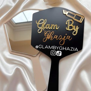 May include: A black hexagonal handheld mirror featuring gold-colored raised text that reads Glam By Ghazia, a gold-colored lipstick icon, and the white text @GLAMBYGHAZIA with Instagram and TikTok icons. A second plain beige handheld mirror is visible.