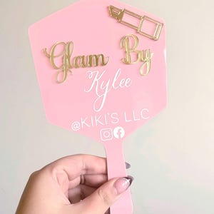 Może przedstawiać: R&oacute;żowe lusterko ręczne ze złotą ikoną szminki i złotym napisem Glam By Kylee. Poniżej znajduje się biały napis @KIKI'S LLC, kt&oacute;remu towarzyszą białe ikony medi&oacute;w społecznościowych Instagram i Facebook u dołu lusterka.