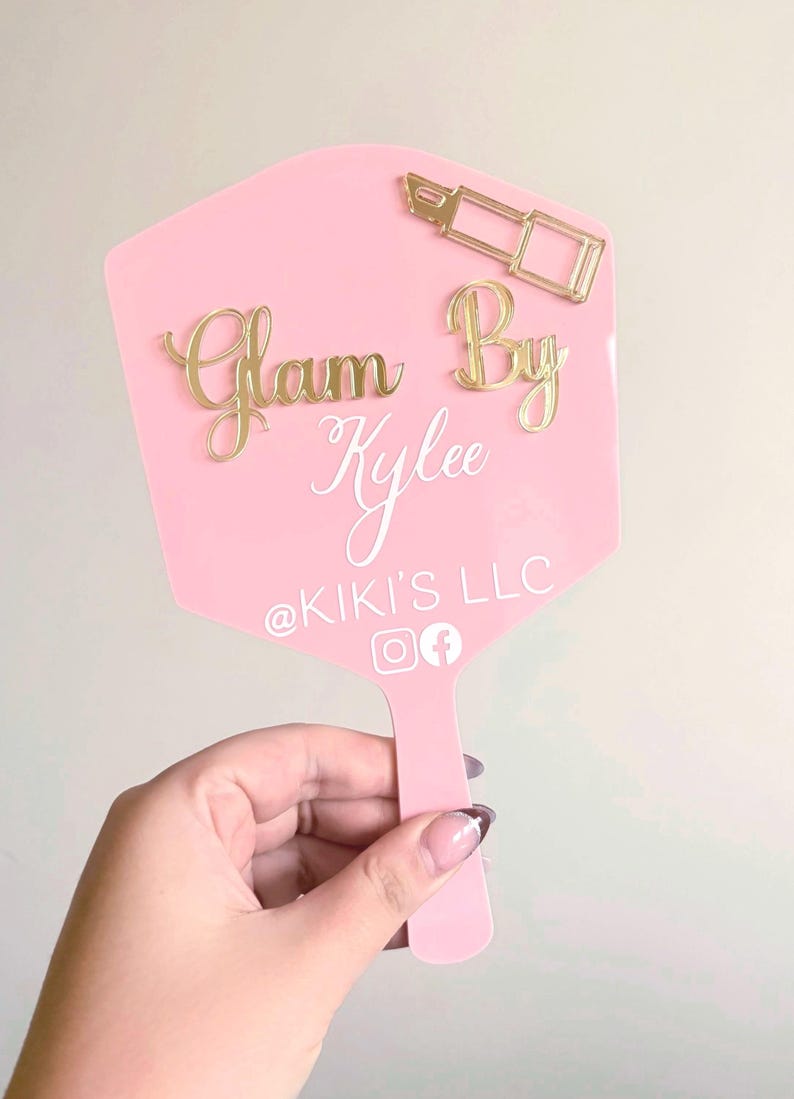 Może przedstawiać: R&oacute;żowe lusterko ręczne ze złotą ikoną szminki i złotym napisem Glam By Kylee. Poniżej znajduje się biały napis @KIKI'S LLC, kt&oacute;remu towarzyszą białe ikony medi&oacute;w społecznościowych Instagram i Facebook u dołu lusterka.