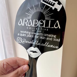 May include: A black handheld mirror featuring a white graphic of a syringe, a sunburst, and a pair of lips. The text reads: ARABELLA COLLECTIVE EST 2020, working towards creating a quiet place for skin and soul, and the Instagram handle @arabella.collective.