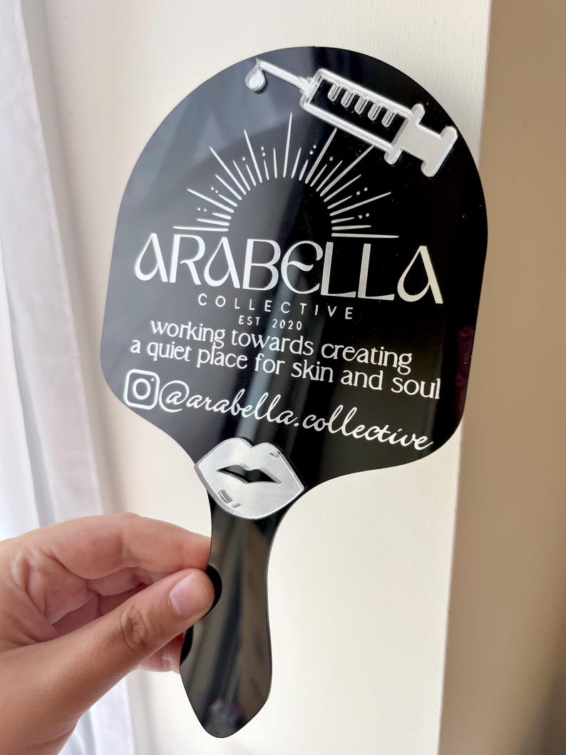 May include: A black handheld mirror featuring a white graphic of a syringe, a sunburst, and a pair of lips. The text reads: ARABELLA COLLECTIVE EST 2020, working towards creating a quiet place for skin and soul, and the Instagram handle @arabella.collective.
