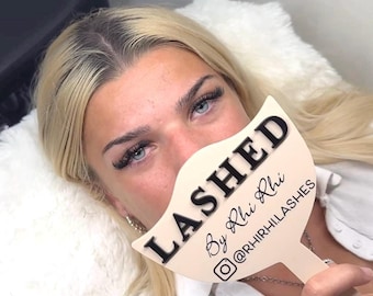 May include: A cream-coloured, fan-shaped handheld mirror featuring the black text LASHED, By Rhi Rhi, and the Instagram handle @RHIRHI.LASHES. The mirror has a handle at the bottom and is held in front of the face.