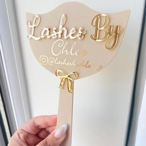 May include: A beige handheld sign with a fan-shaped top and a handle. The sign features the text "Lashes By Chloe" in raised gold lettering, a gold Instagram icon, the handle "@lashesbychloe_c", and a small gold bow accent at the base.