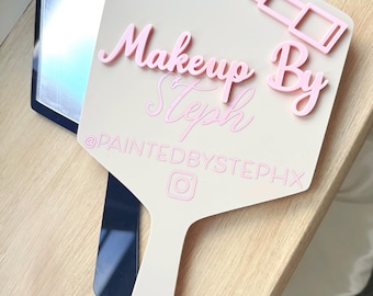 Könnte beinhalten: Ein beigefarbener Handspiegel mit einer erhabenen rosa Schrift mit der Aufschrift Makeup By Steph, einem rosa Lippenstift-Symbol und dem Text @PAINTEDBYSTEPHX mit einem Instagram-Logo. Der Spiegel ist sechseckig mit Griff und dunkelblauer Spiegelfläche.