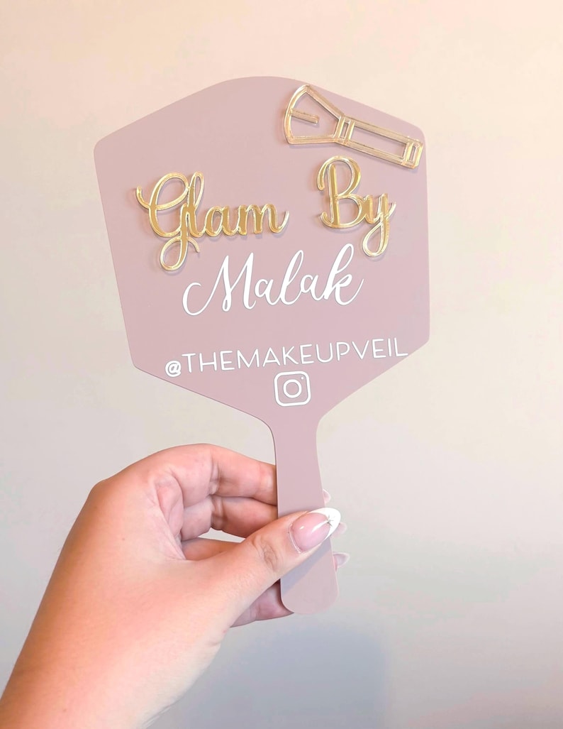 Może przedstawiać: Sześciokątne lusterko ręczne w kolorze przybrudzonego r&oacute;żu ze złotymi akrylowymi literami z napisem Glam By Malak, złotą ikoną pędzla do makijażu oraz białym napisem @THEMAKEUPVEIL nad białą ikoną logo Instagrama.