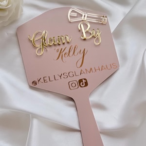Może przedstawiać: Pudrowo-r&oacute;żowe lusterko ręczne ze złotym napisem z akrylu Glam By Kelly, tekstem @KELLYSGLAM.HAUS oraz ikonami medi&oacute;w społecznościowych. Ikona pędzla do makijażu z przezroczystego akrylu jest przymocowana w prawym g&oacute;rnym rogu powierzchni lusterka.