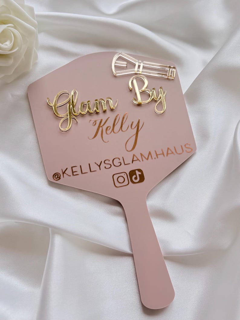 Może przedstawiać: Pudrowo-r&oacute;żowe lusterko ręczne ze złotym napisem z akrylu Glam By Kelly, tekstem @KELLYSGLAM.HAUS oraz ikonami medi&oacute;w społecznościowych. Ikona pędzla do makijażu z przezroczystego akrylu jest przymocowana w prawym g&oacute;rnym rogu powierzchni lusterka.