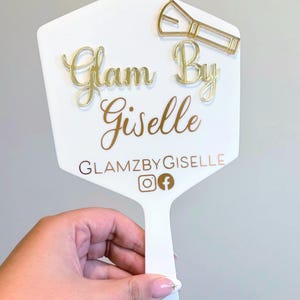 Może przedstawiać: Białe lusterko ręczne z wypukłym złotym napisem Glam By Giselle. Nad tekstem znajduje się złota ikona pędzla do makijażu. Pod nazwą widnieje napis GLAMZBYGISELLE wraz z ikonami serwis&oacute;w społecznościowych Instagram i Facebook.
