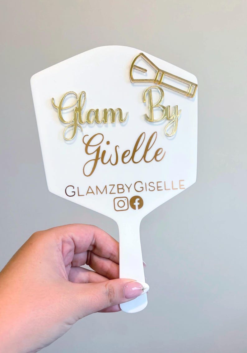 Może przedstawiać: Białe lusterko ręczne z wypukłym złotym napisem Glam By Giselle. Nad tekstem znajduje się złota ikona pędzla do makijażu. Pod nazwą widnieje napis GLAMZBYGISELLE wraz z ikonami serwis&oacute;w społecznościowych Instagram i Facebook.