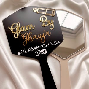 Może przedstawiać: Czarne lusterko ręczne ze złotą ikoną szminki i złotym napisem Glam By Ghazia. Pod tekstem znajduje się biały uchwyt @GLAMBYGHAZIA z ikonami Instagrama i TikToka. Drugie beżowe lusterko ręczne jest częściowo widoczne za czarnym lusterkiem.