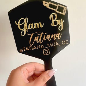 Może przedstawiać: Czarne lusterko ręczne o sześciokątnym kształcie ze złotym napisem Glam By Tatiana, uchwytem @TATIANA_MUA_OC, ikoną Instagrama oraz złotą grafiką szminki umieszczoną w prawym g&oacute;rnym rogu powierzchni lustra.